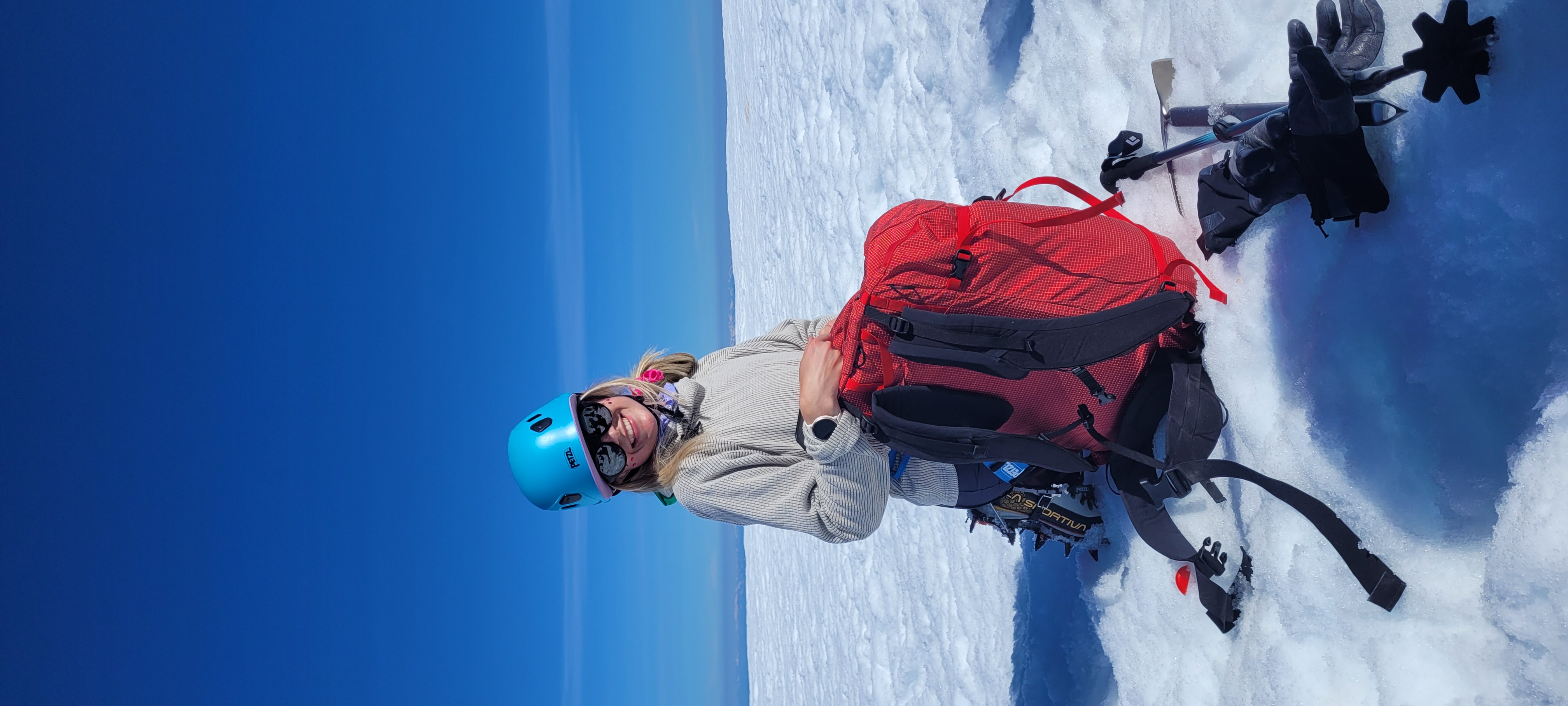 Saachi at the summit, smiling, blue helmet, red pack, pure blue sky behind