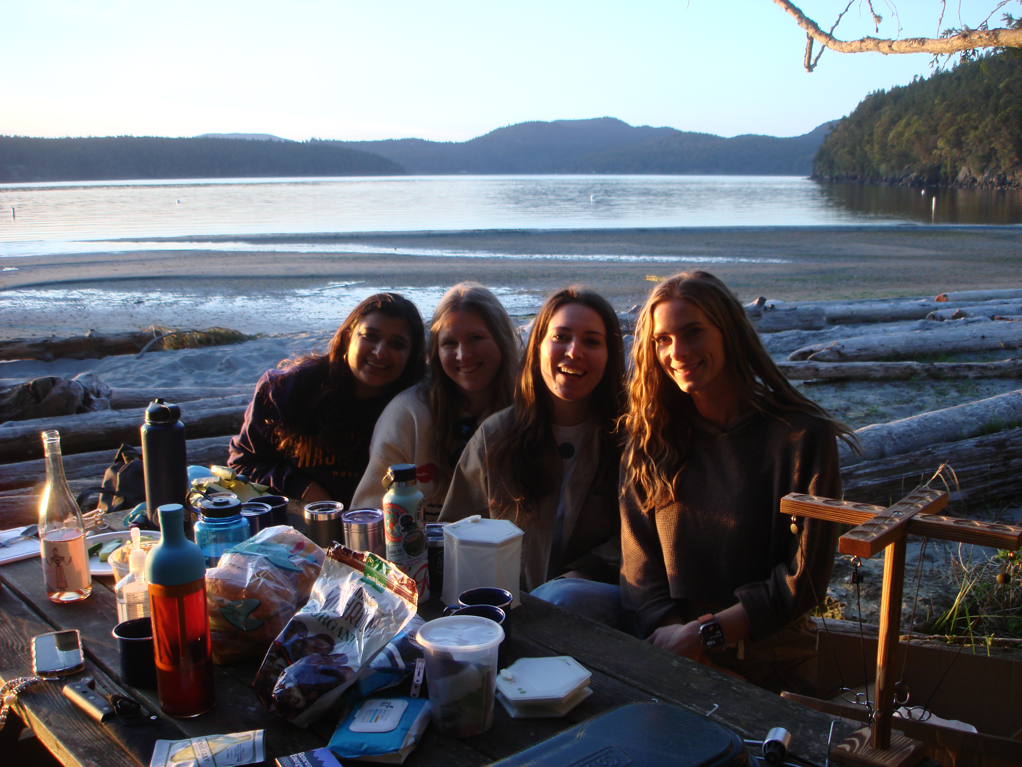 Four of us at a picnic table on the beach at golden hour, water and mountains behind