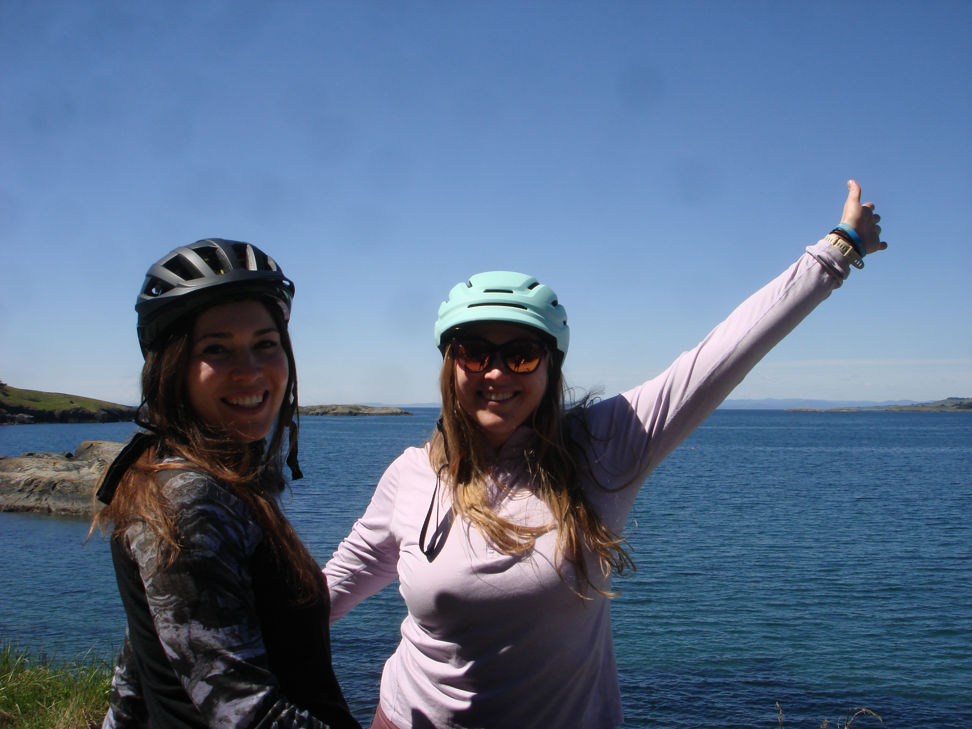 Two of us in bike helmets at a scenic water overlook, thumbs up, deep blue water behind